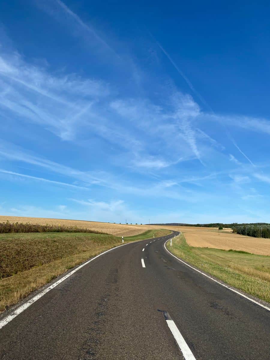 gray asphalt road between green grass field under blue and white sunny cloudy sky during daytime